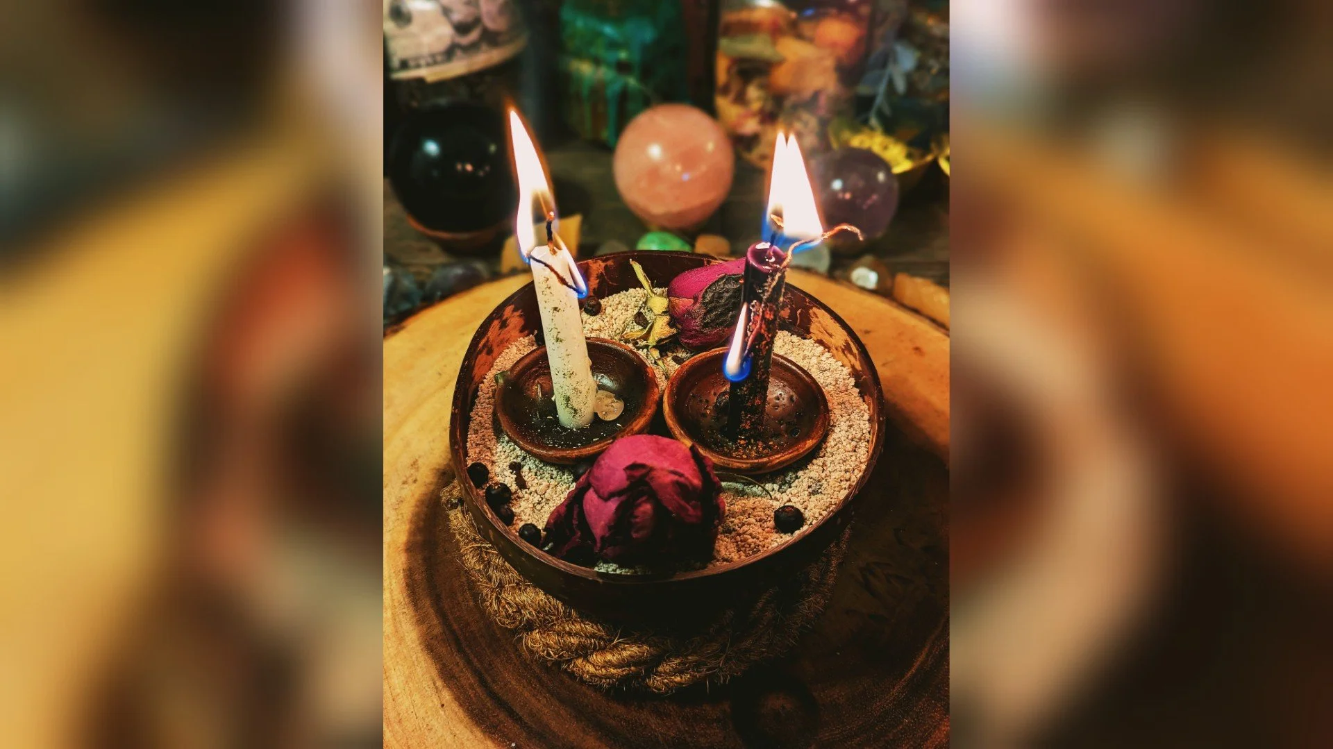 A decorative arrangement with three lit candles, dried flowers, and black peppercorns in a bowl, surrounded by crystals and beads on a wooden surface.