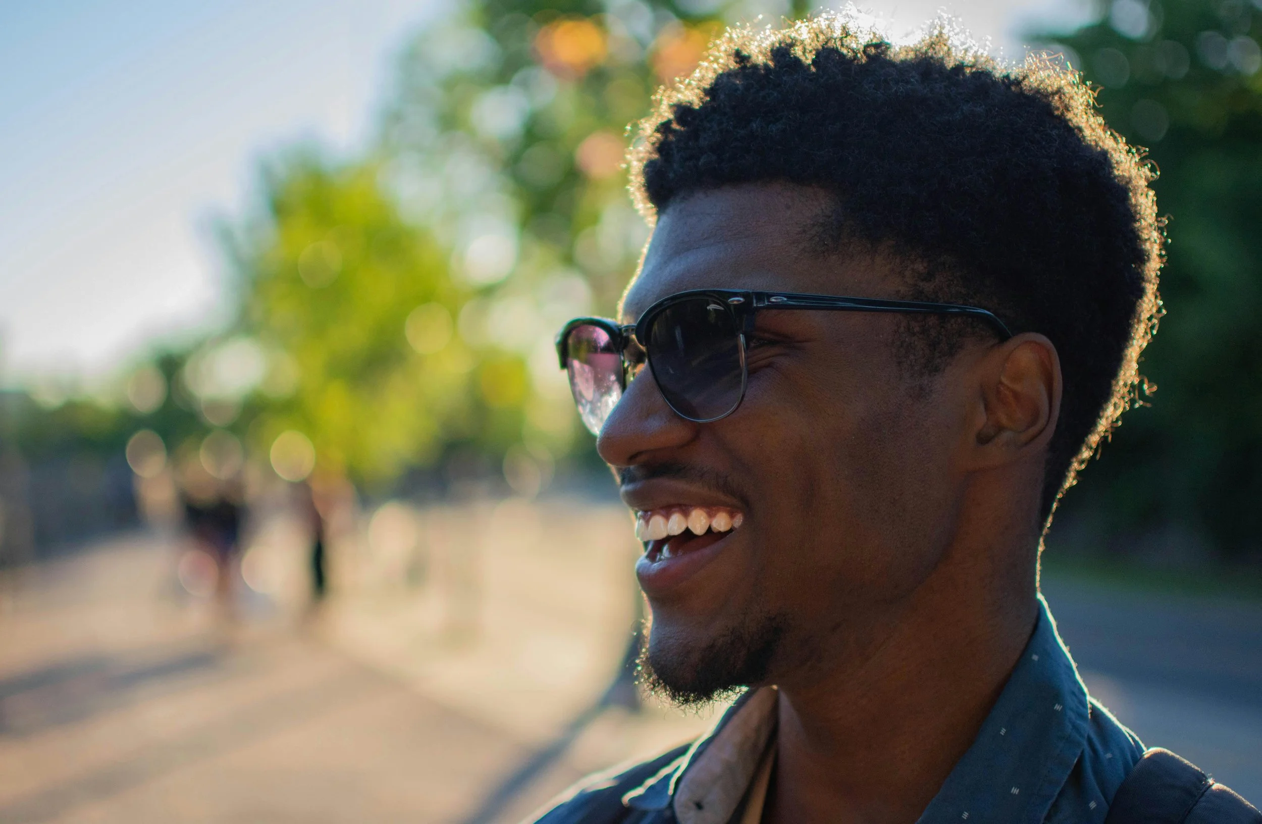 A young man with dark skin, short curly hair, wearing sunglasses and a blue shirt, smiling outdoors during sunset.