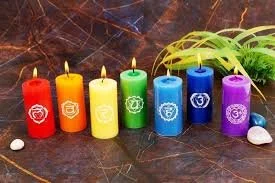 Seven candles in red, orange, yellow, green, blue, purple, and violet, each with a different symbol, placed on a wooden surface with small white stones and green plants in the background.