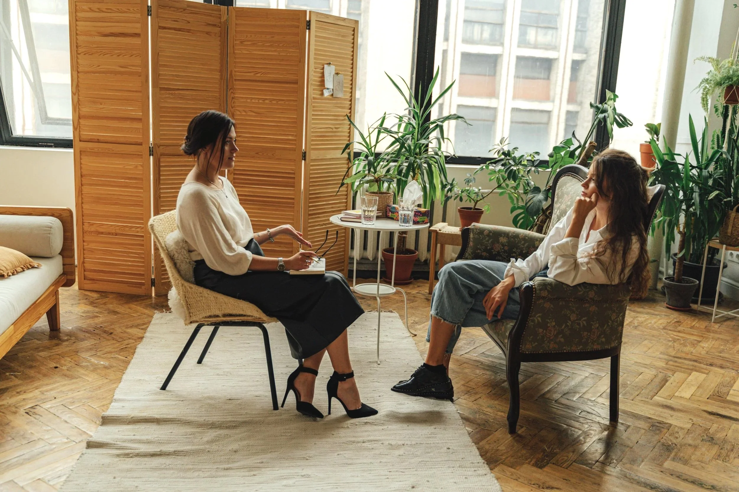 Two women having a conversation in a cozy, plant-filled room with large windows, wooden flooring, and a folding room divider.