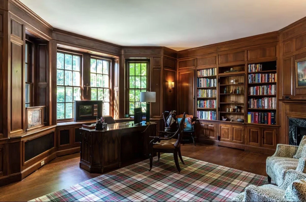 A wood-paneled home office with a large desk near windows, a bookshelf filled with books, and a plaid area rug.