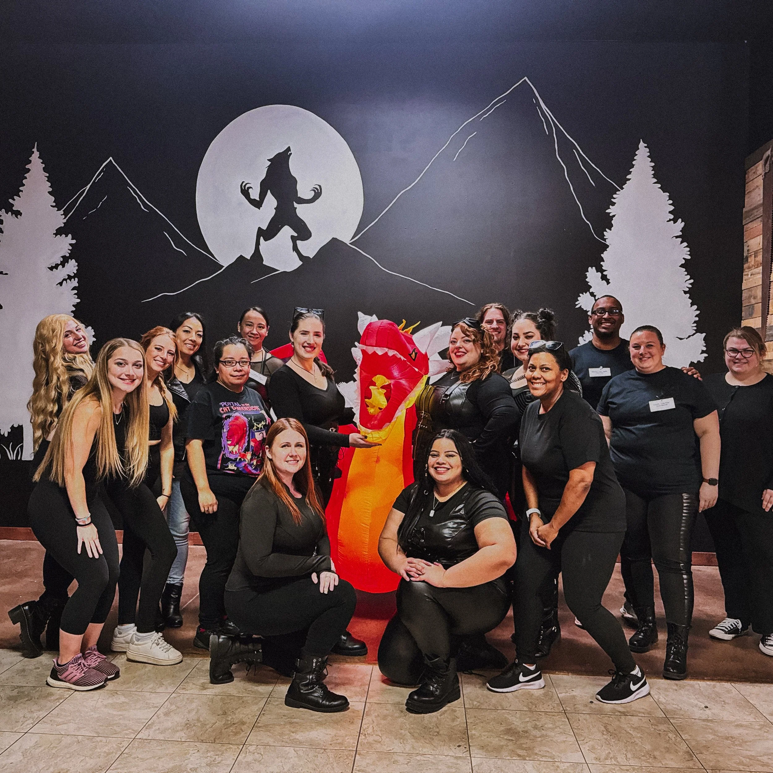 Group of people posing with a red dragon costume in front of a mural featuring a werewolf howling at a full moon, with mountains and trees in the background.