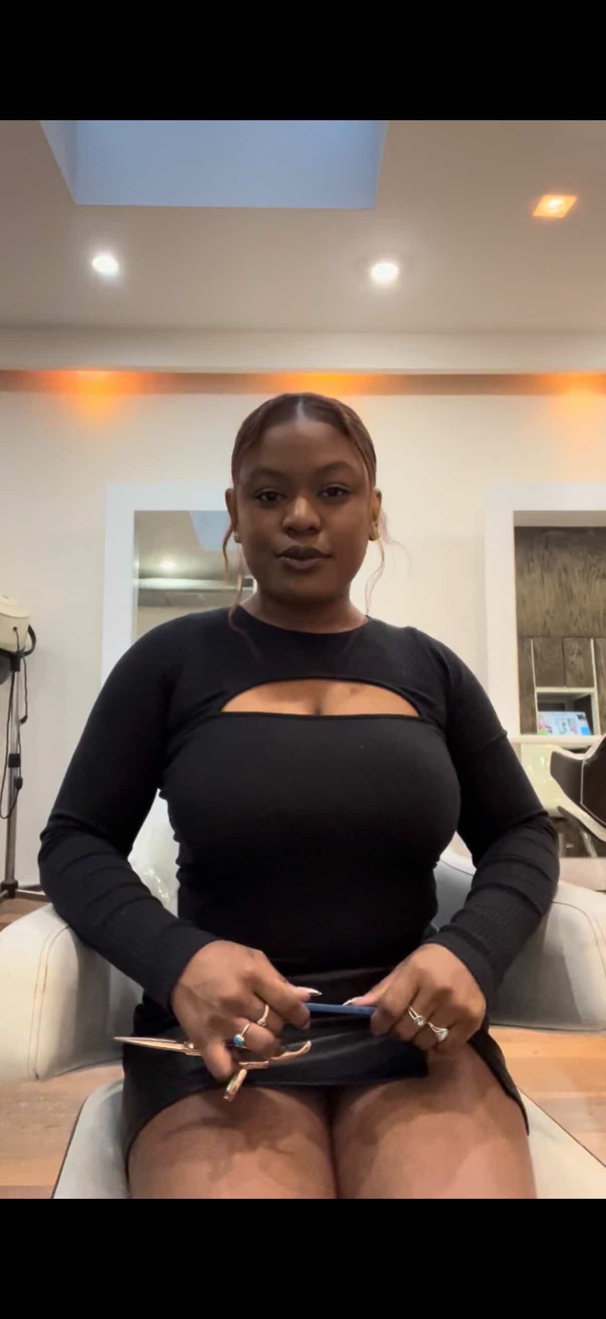 A woman with dark hair sitting indoors in a black long-sleeve top with a cut-out, holding a smartphone, in a room with beige walls and ceiling, with lights and mirrors in the background.