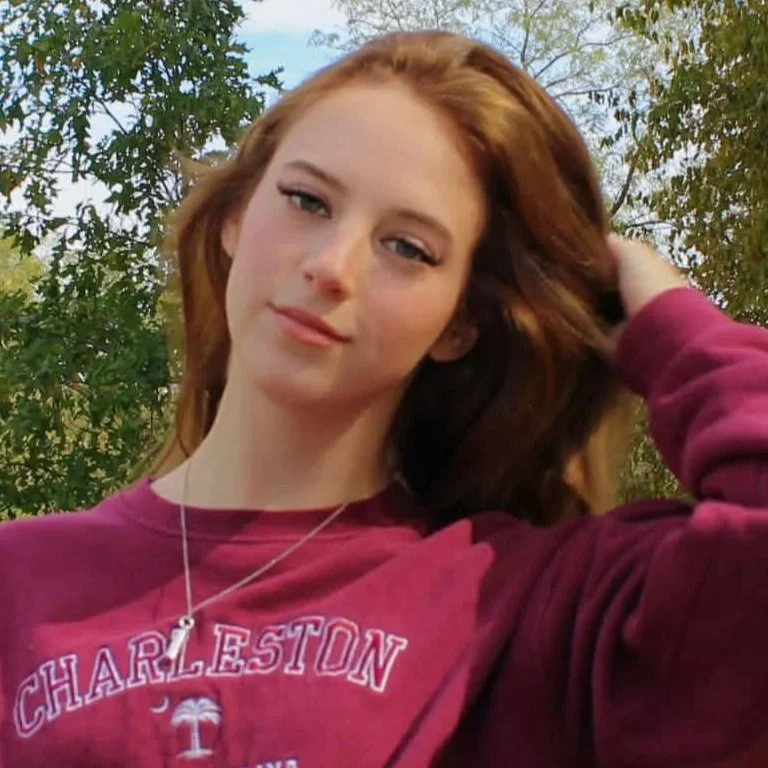 A young woman with red hair, wearing a maroon Charleston sweatshirt and a necklace, is outdoors with trees in the background, looking at the camera with a slight smile.