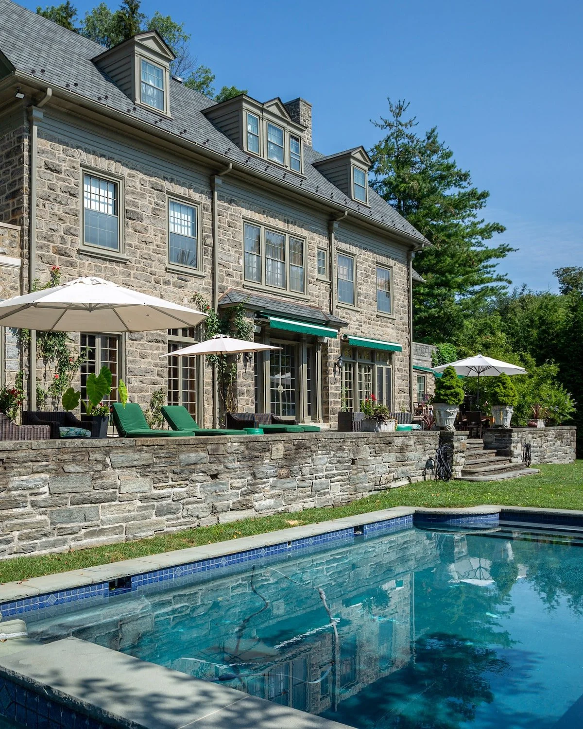 The image shows the backyard of a stone house with multiple windows, a patio area with umbrellas and sun loungers, and a swimming pool in the foreground under a clear blue sky.