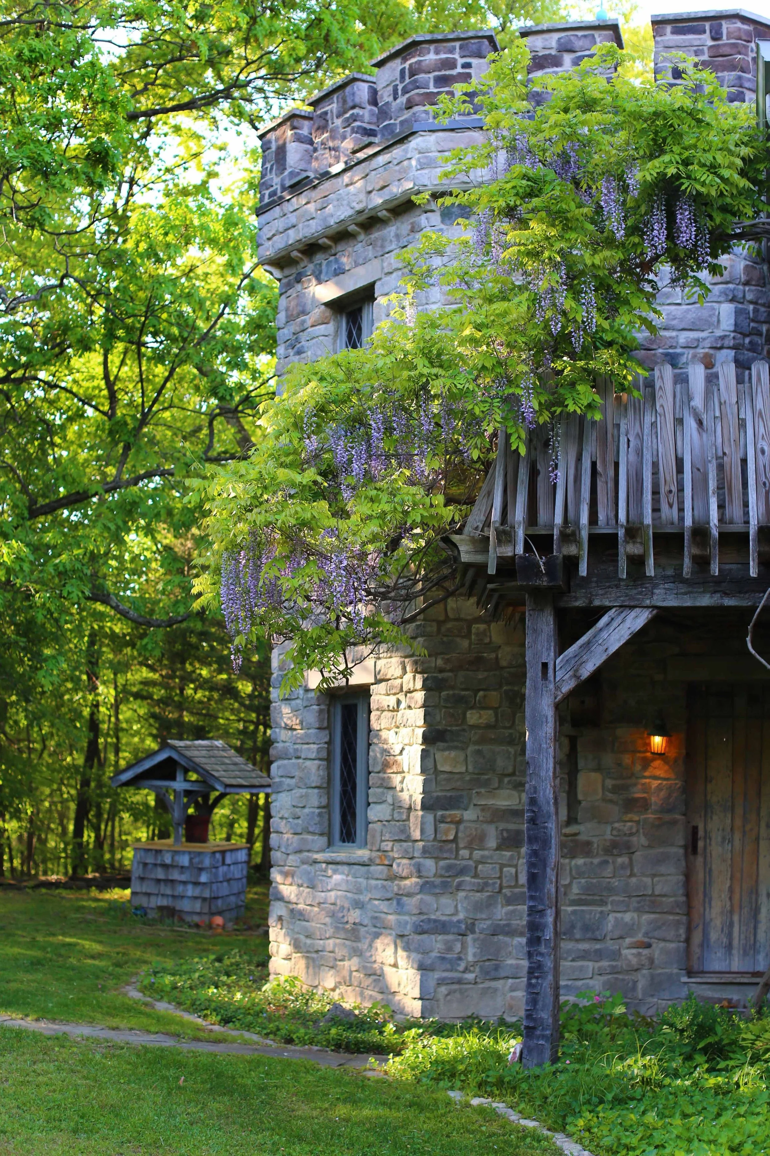 A stone building with a wooden balcony and a small well in the yard surrounded by green trees and grass, with flowering vines on the balcony.