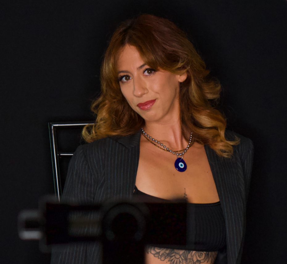 A woman with red hair, wearing a black pinstripe blazer and a black top, sitting against a black background, posing for a photo.