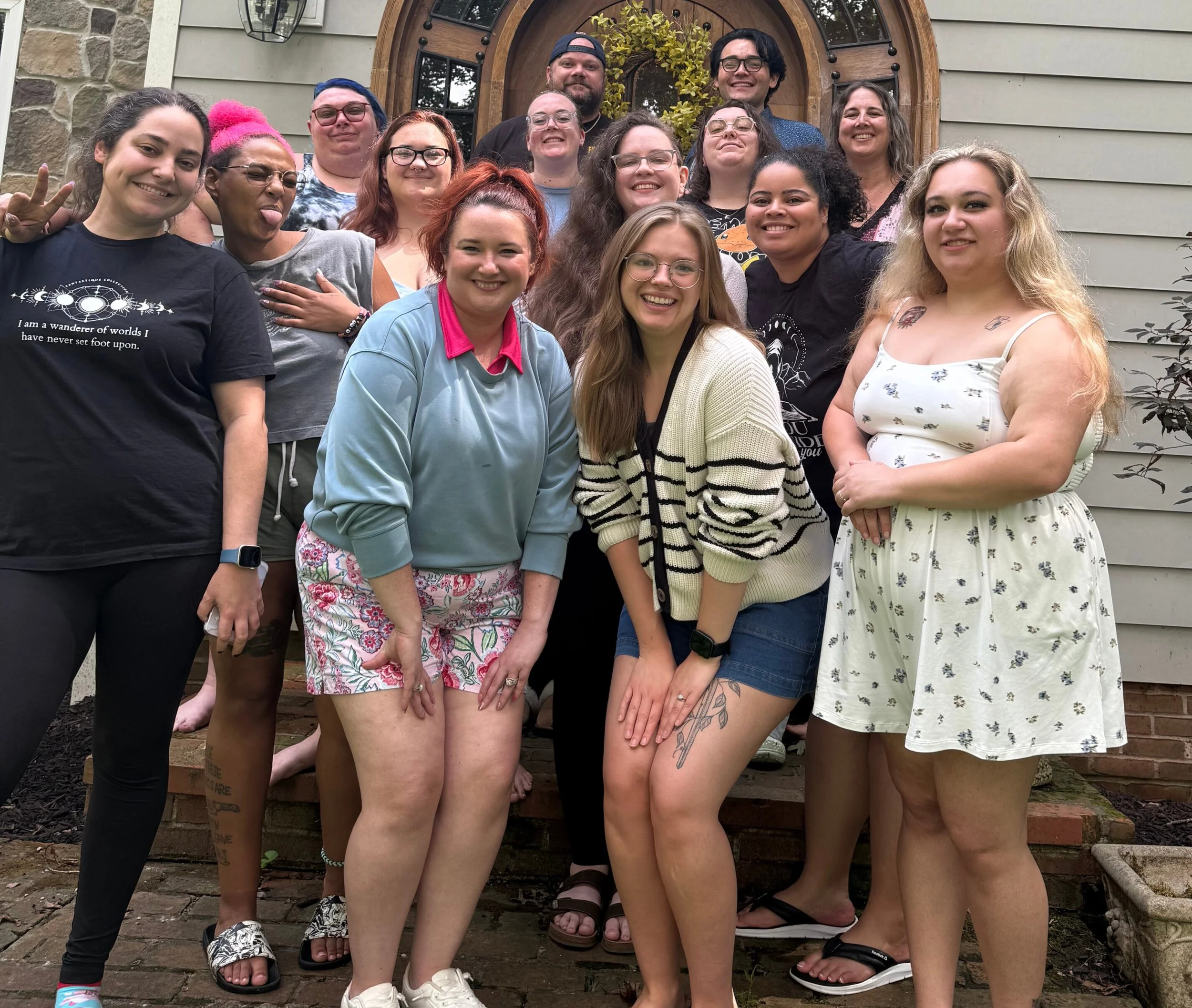 Group of people posing outside in front of a house with a wooden door and stone accents, smiling and making various gestures.