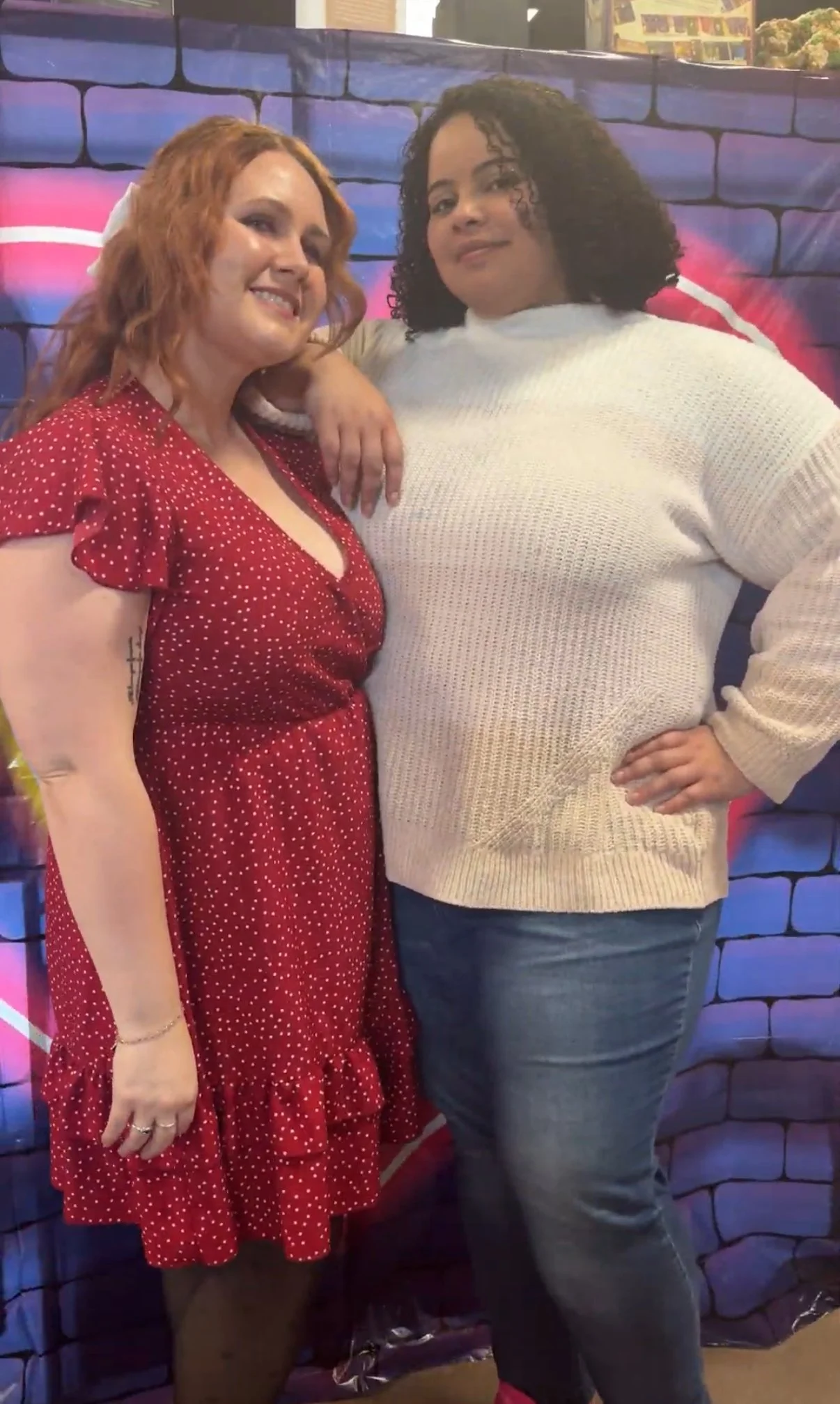 Two women standing close together in front of a colorful, painted brick wall, one with red hair and a red polka-dot dress, the other with dark curly hair and a cream sweater.