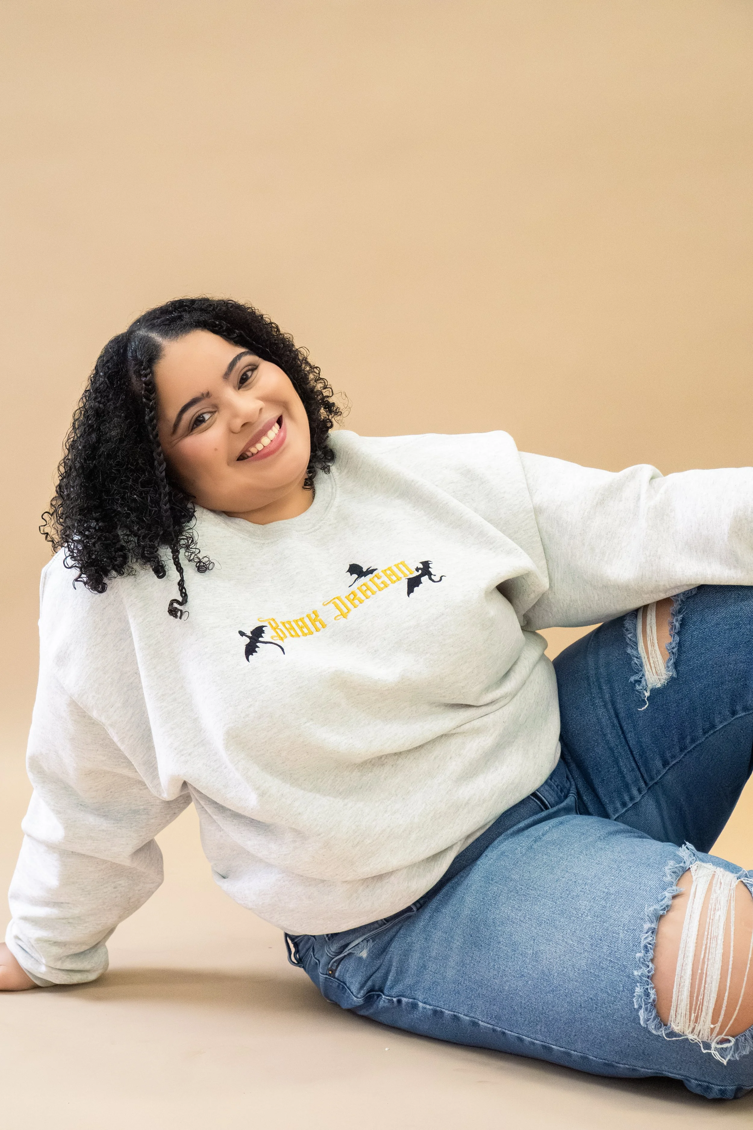 Young woman with curly black hair smiling, wearing a gray sweatshirt with Halloween-themed text and images, sitting on the floor against a beige background.
