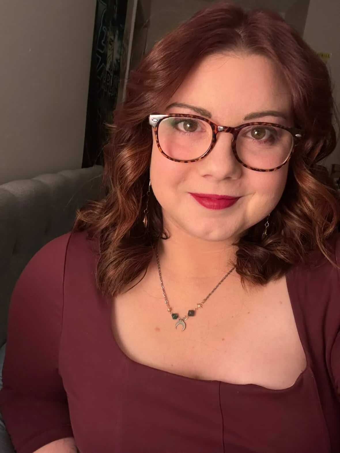 A woman with reddish-brown, wavy hair and glasses, wearing a maroon top and a necklace with a crescent moon pendant, smiling and sitting on a gray couch in a room with a dark background.