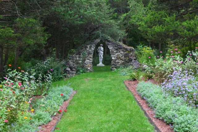 Garden with a stone archway leading to a statue of a woman, surrounded by colorful flowers and trees.