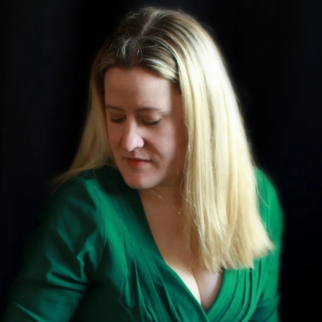 A woman with blonde hair wearing a green top, looking downward against a dark background.