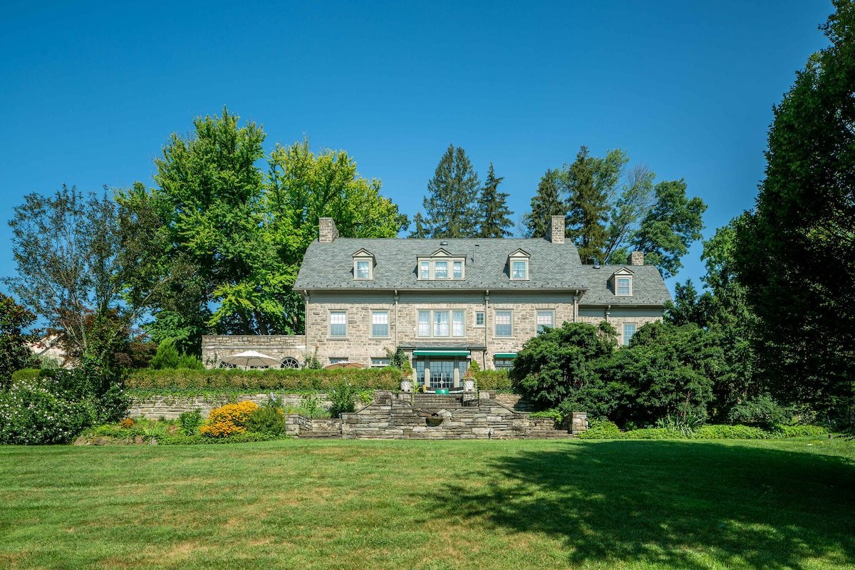 A large stone mansion with multiple chimneys and dormer windows, surrounded by tall trees and a well-manicured lawn, under a clear blue sky.
