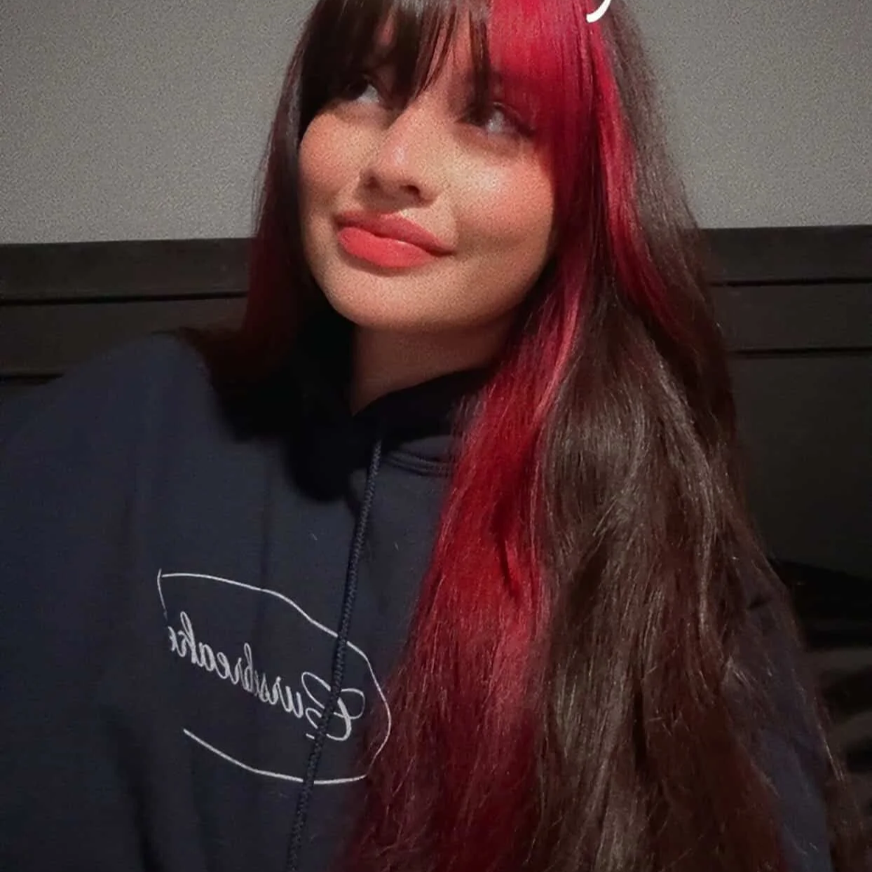 A young woman with long brown hair and red highlights, wearing a black hoodie, smiling softly while looking to her left.