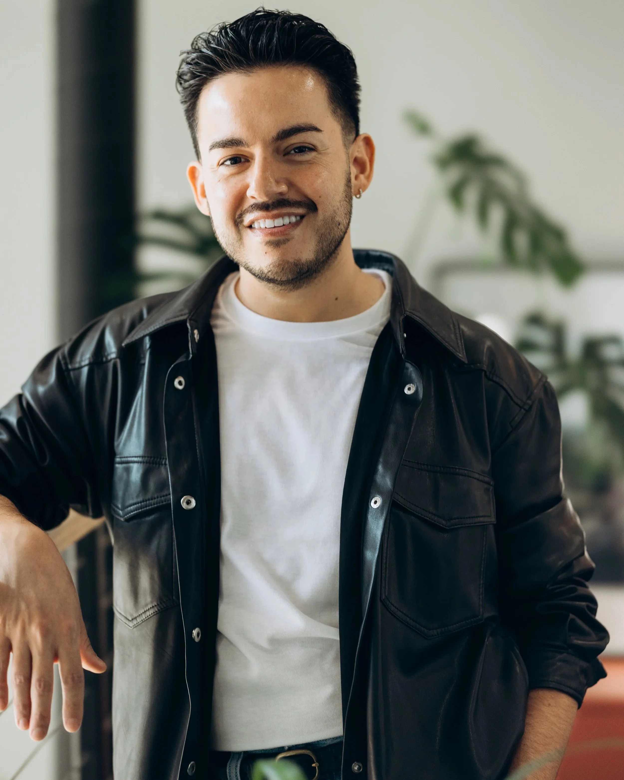 A young man with dark hair, a beard, and earrings, smiling and posing indoors next to a plant.