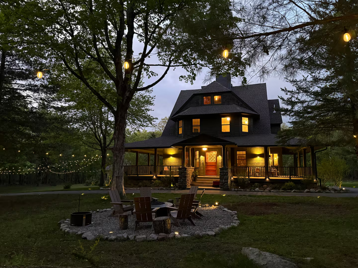 A large, dark-colored house with warm interior lighting, surrounded by trees and string lights, with a gravel fire pit area and outdoor seating in the foreground during early evening.