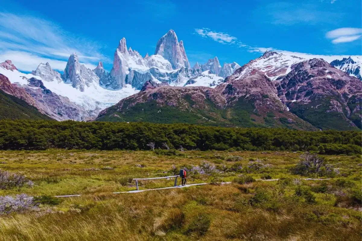 A person hiking on a trail in a valley with green grass and shrubs, with tall snow-capped mountains and blue sky in the background.