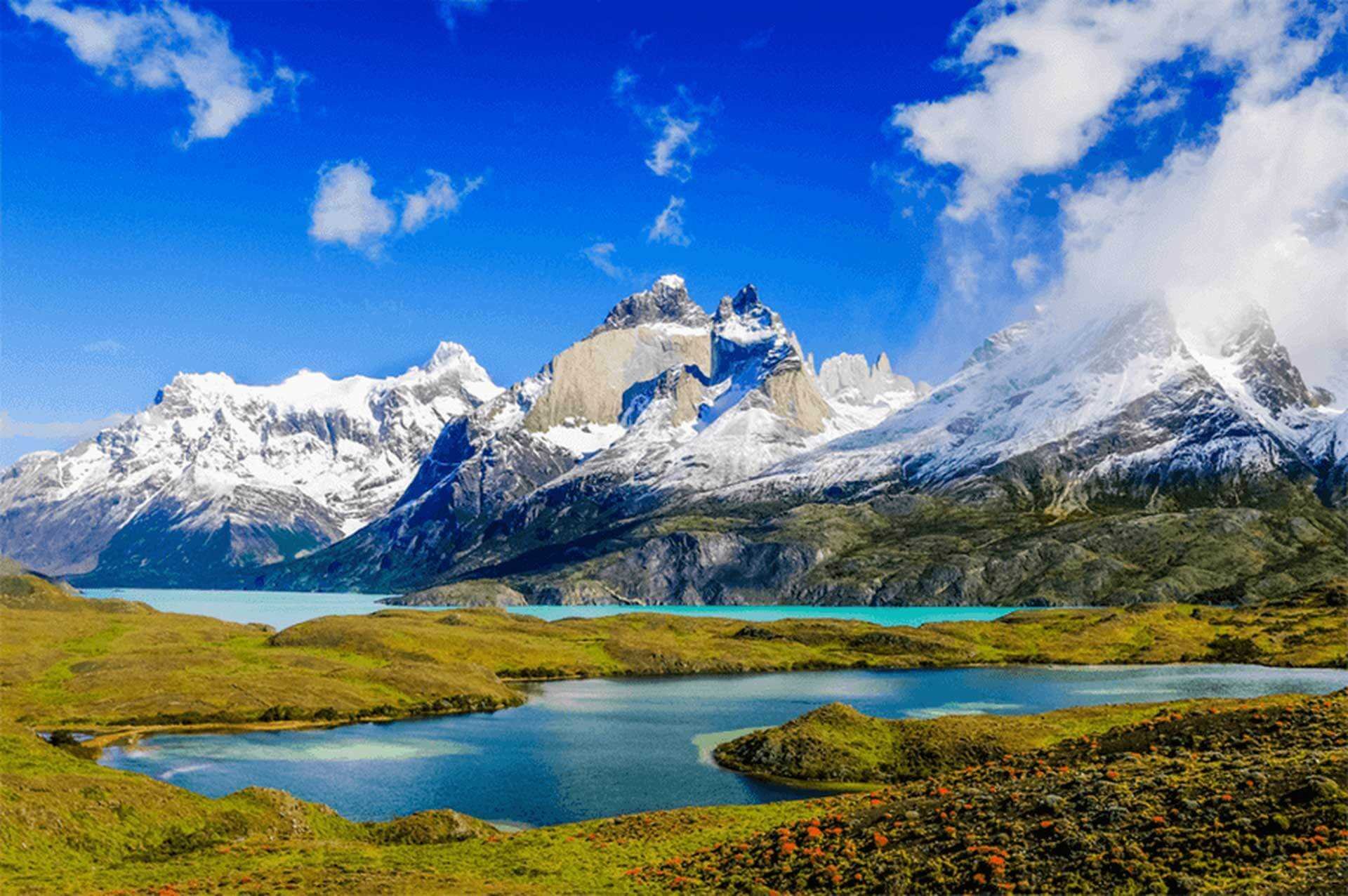 Snow-capped mountains with a blue lake in the foreground, green rolling hills, and a bright blue sky with scattered clouds.