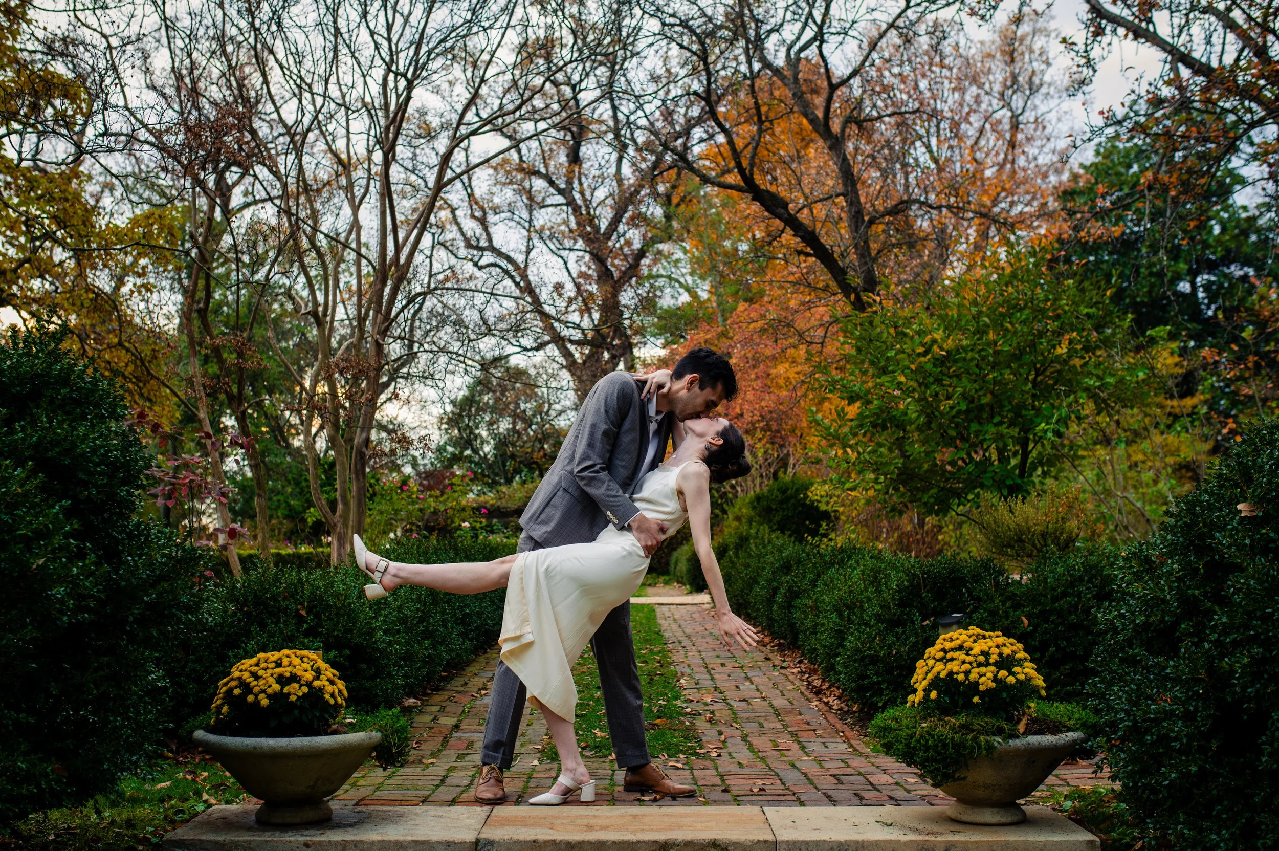 A couple dressed in formal attire sharing a kiss in a garden during autumn with colorful trees and flower pots.
