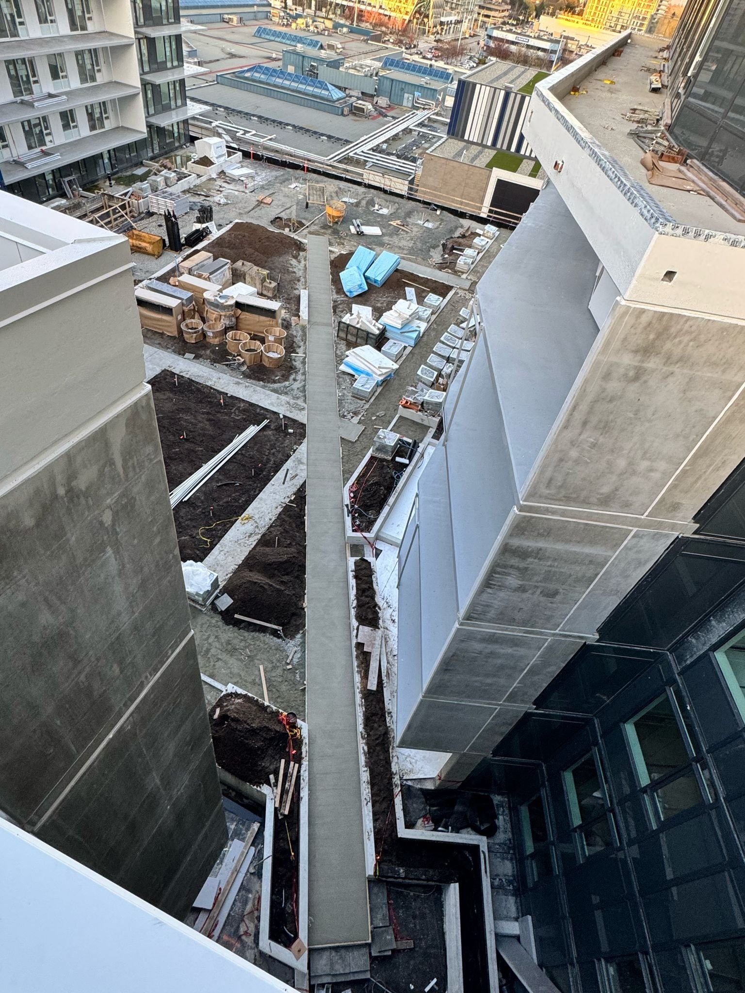 A bird's eye view of a construction site on a high-rise building's balcony, with dirt, construction materials, and equipment scattered, and a cityscape in the background.
