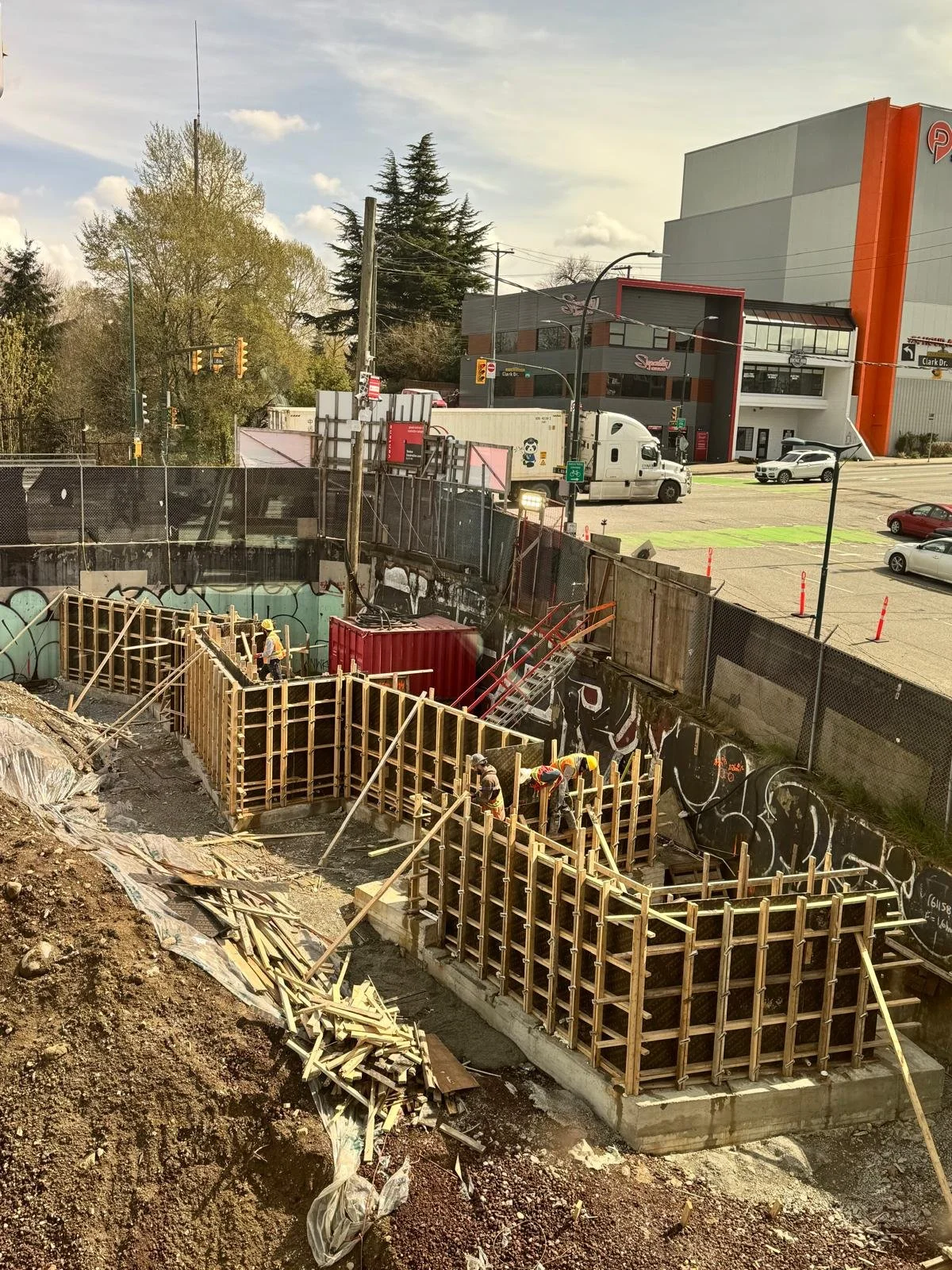 Construction site with workers building wooden formwork for concrete in an urban area near a parking lot and commercial buildings.