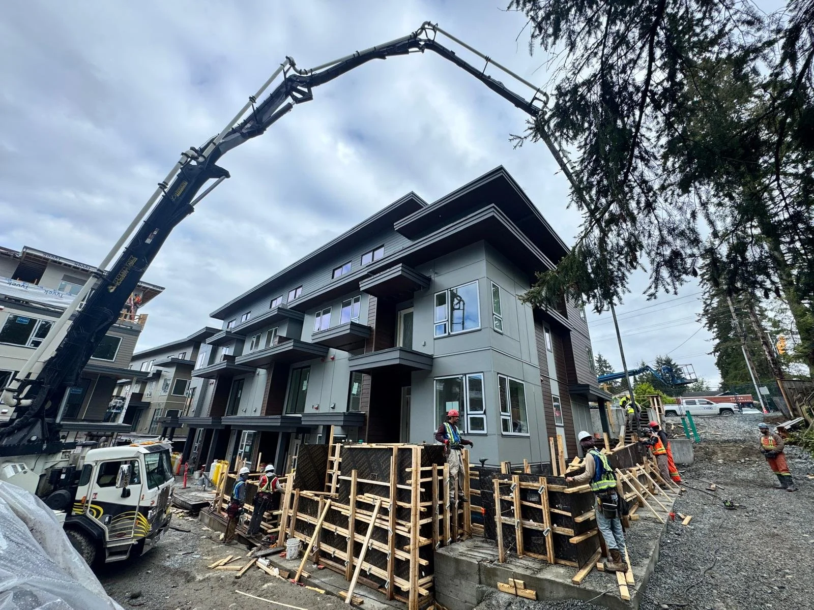 Construction site with workers building a multi-story residential building, wooden scaffolding, and a crane in use, on a cloudy day.