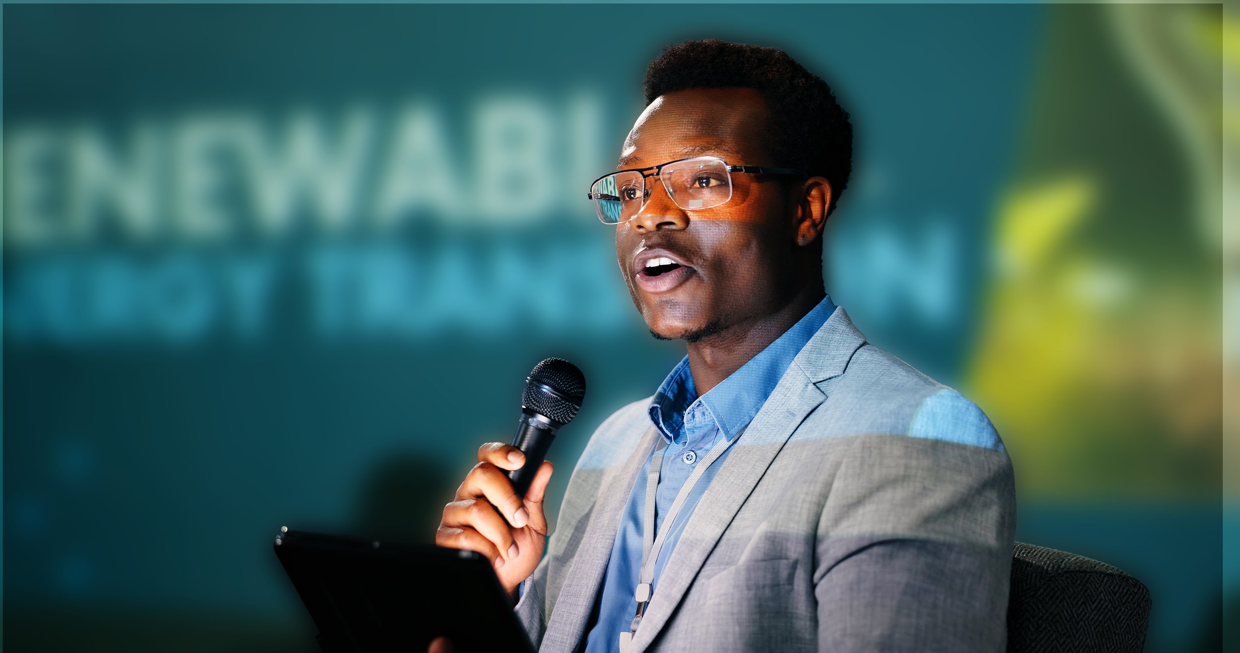 A man wearing glasses and a gray suit is speaking into a microphone while holding a tablet.