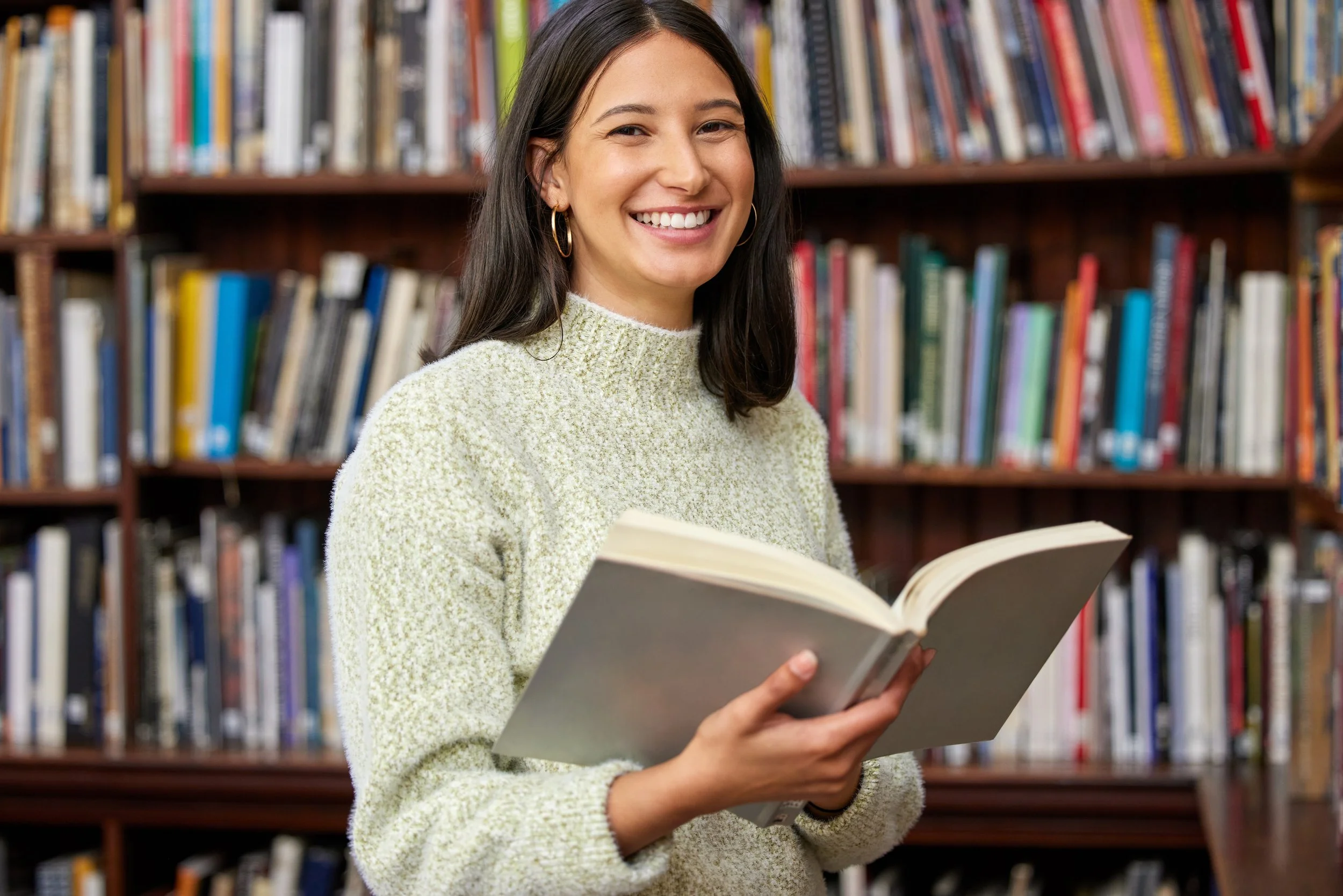 A young woman with dark hair, wearing a light-colored sweater and gold hoop earrings, smiling while holding an open book in a library with rows of bookshelves.