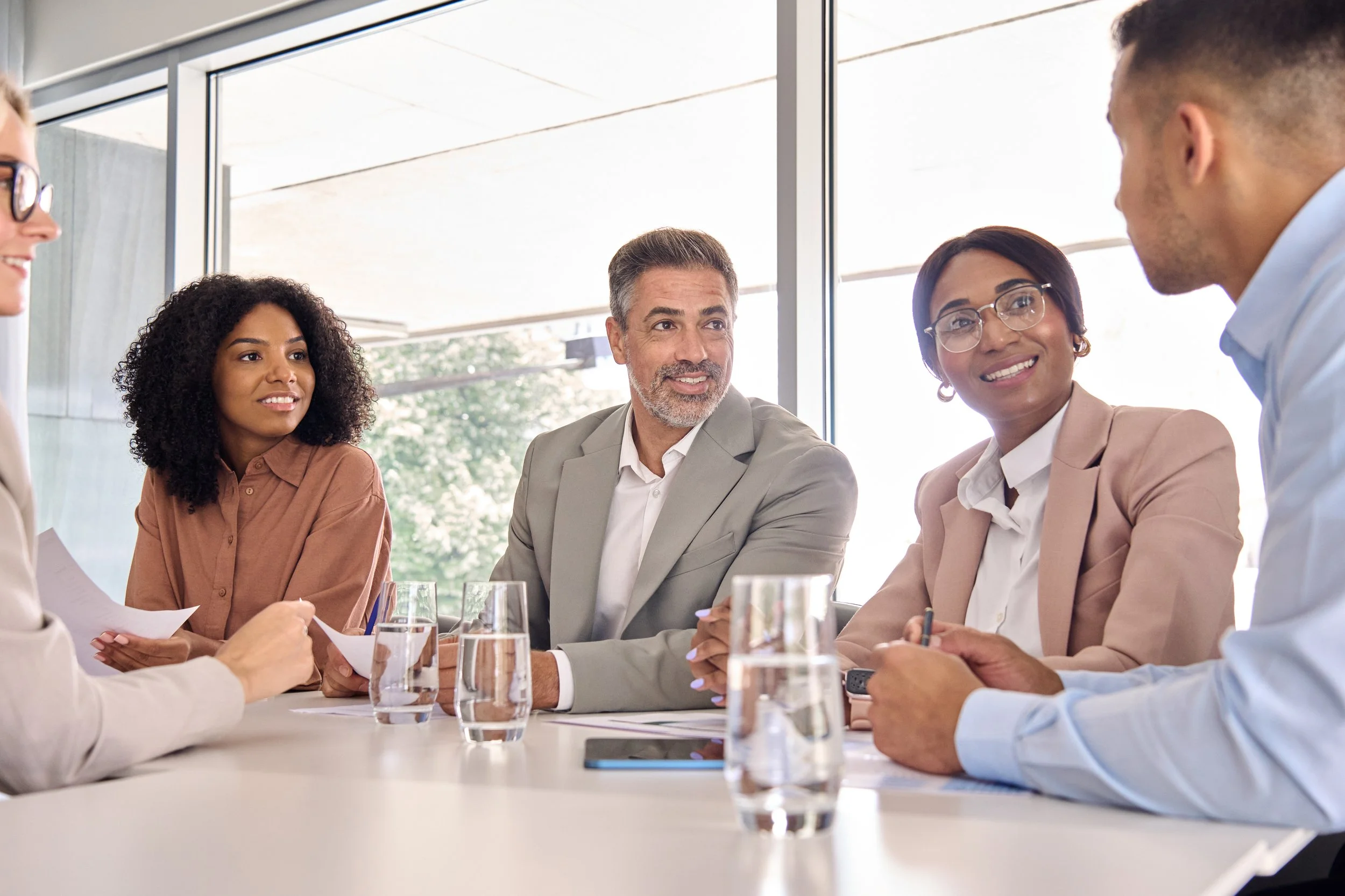 Five diverse professionals are sitting at a table in a meeting room, engaged in a discussion, with glasses of water on the table and large windows in the background.