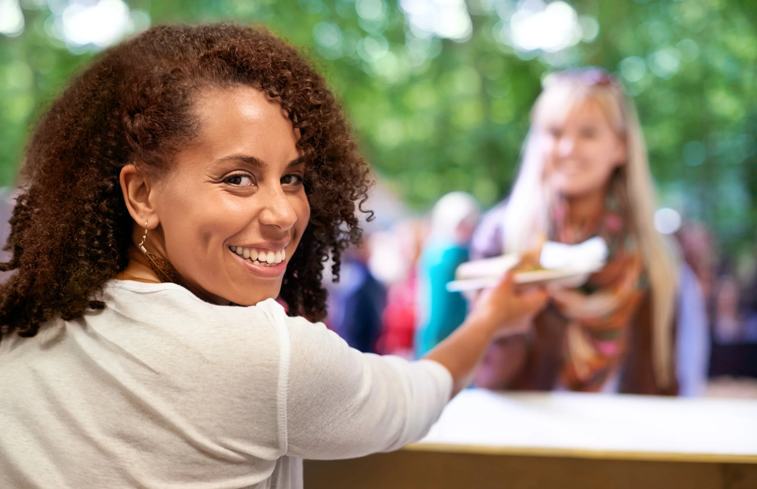 Smiling woman with curly hair reaching for a plate at an outdoor gathering, with blurred group of people in the background.