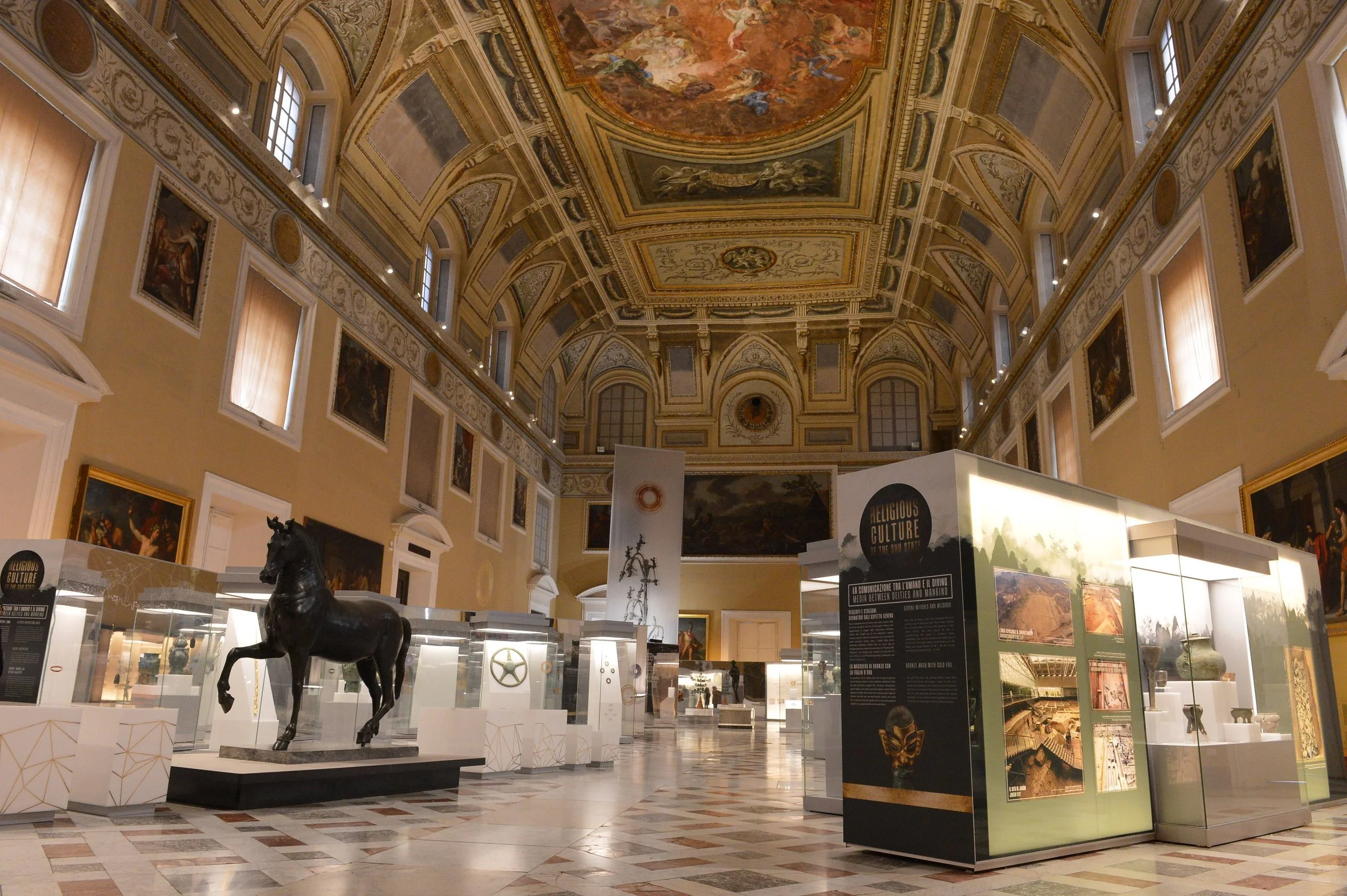 Interior of a grand art museum with ornate ceiling paintings, framed artworks on the walls, and artifacts displayed in glass cases, including a black horse statue in the center.