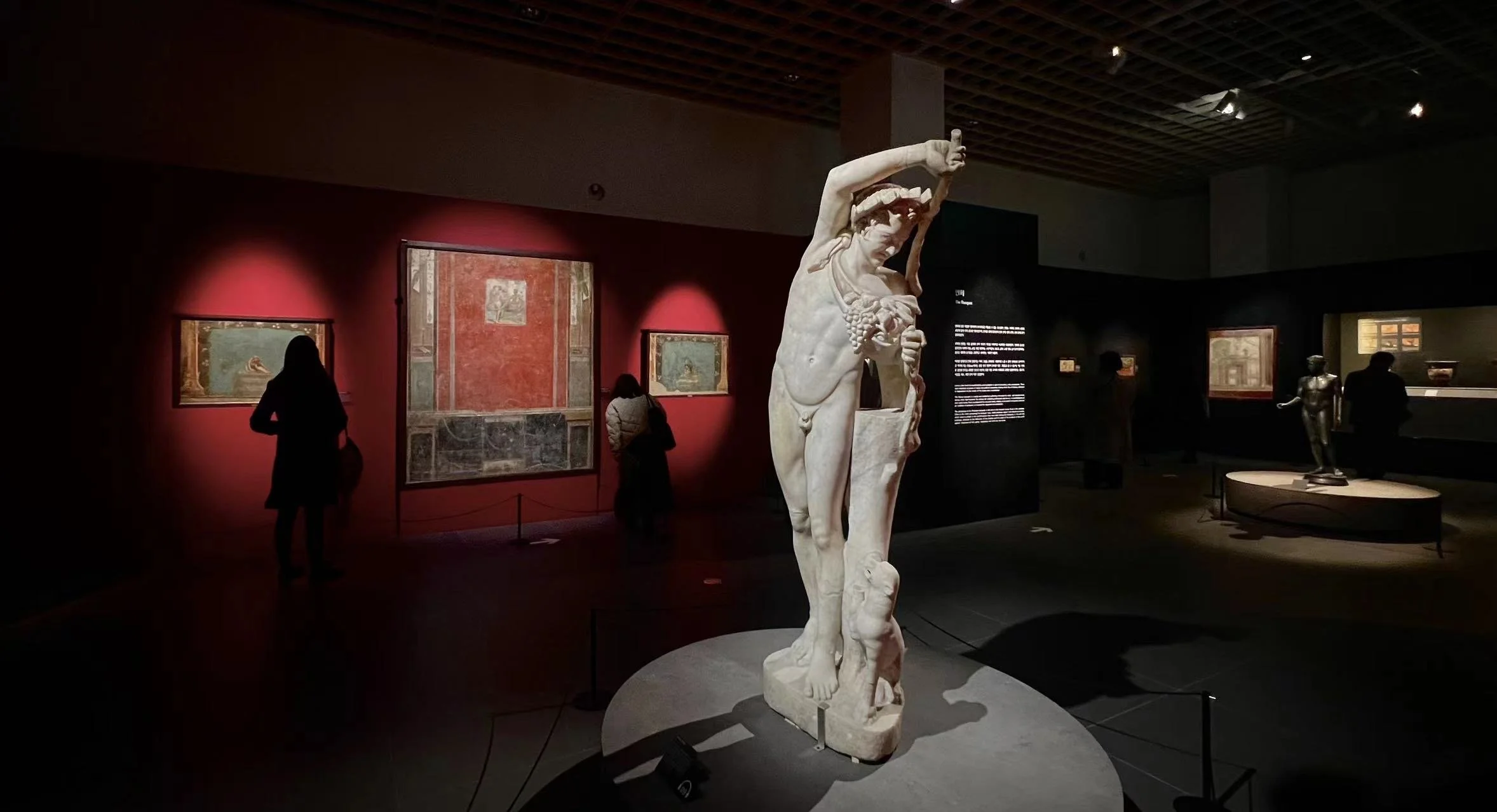 Marble statue of a male figure with an outstretched arm holding an object, displayed in a museum gallery with dim lighting and other artworks in the background, including paintings and a sculpture of a woman.