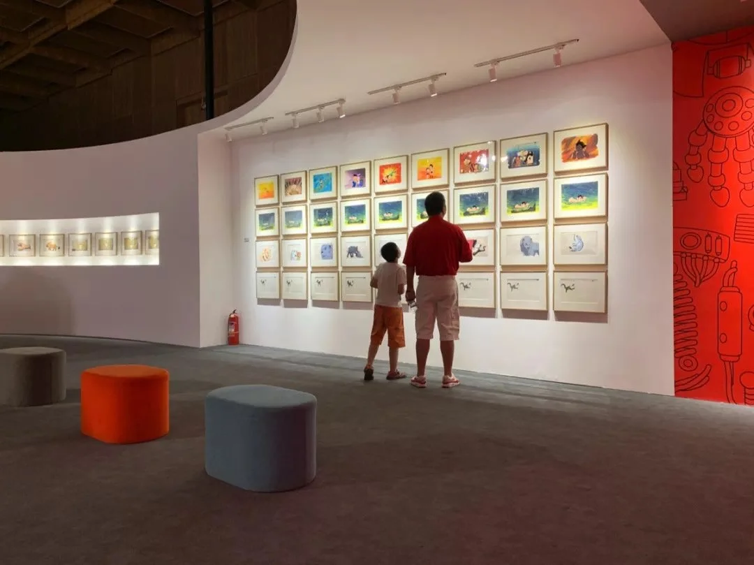 Two people, a child and an adult, looking at colorful artwork displayed on a white wall in an art gallery. The gallery has a curving wall, a gray carpet, and small stools in orange, blue, and beige. There is a fire extinguisher on the floor near the 