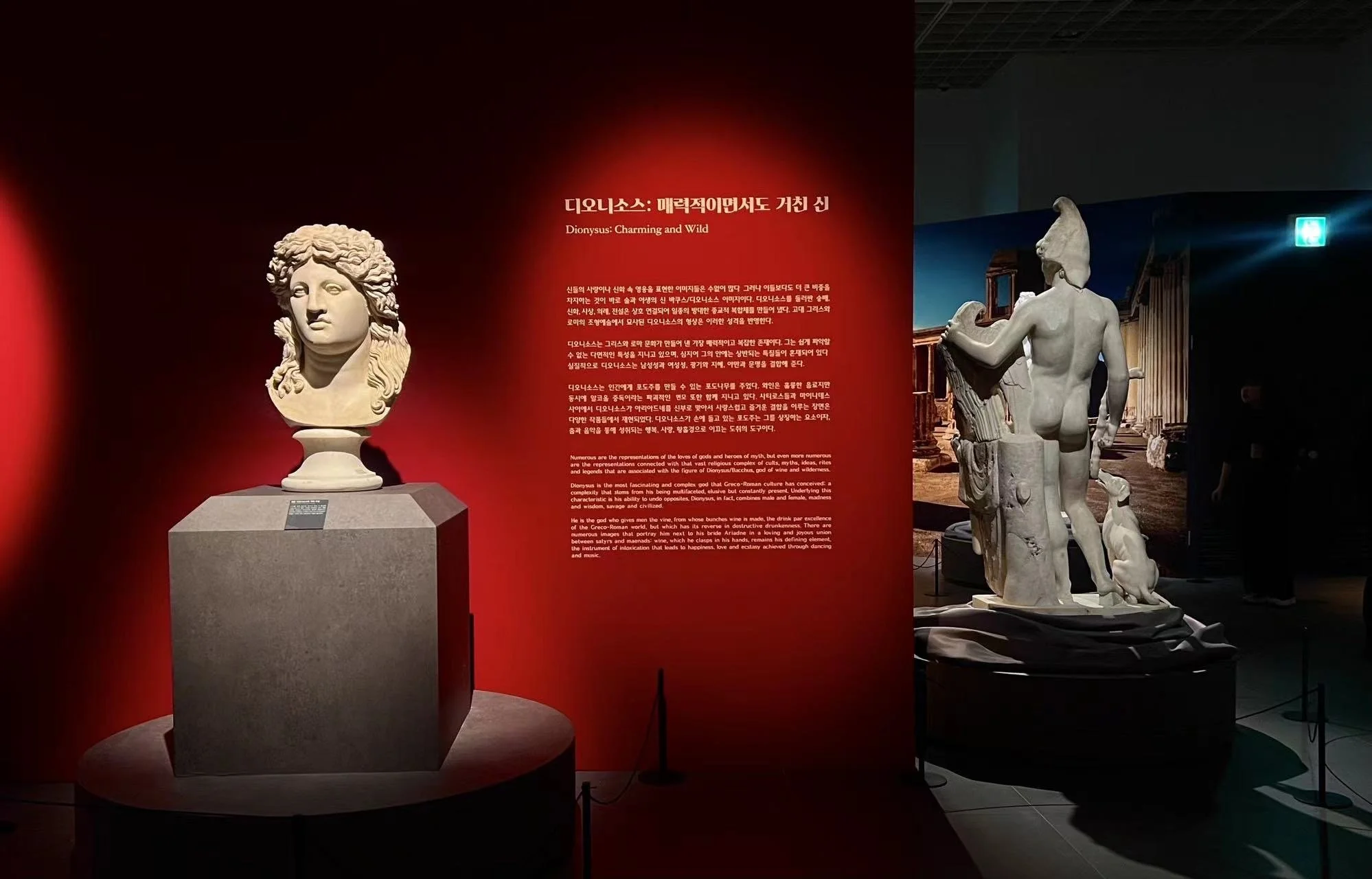 Ancient Greek sculptures displayed in a museum with informational panels; one bust on a pedestal on the left and a full statue of a male figure on the right, with a red wall and dark background.