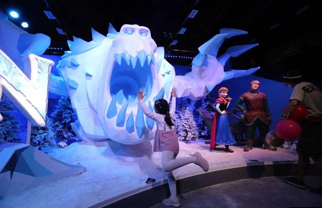 A young girl with long black hair, wearing a pink and white outfit, kneeling with arms raised in front of large snow-themed sculptures of a blue-eyed white wolf and Frozen characters Elsa and Kristoff at an indoor exhibit.