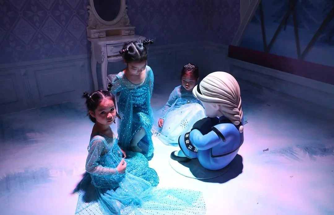 Three young girls in blue dresses and a person in a cartoon character costume sitting on the floor in a room with soft lighting.