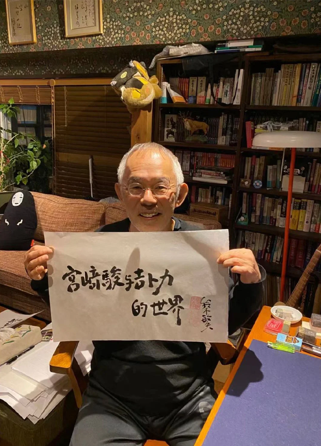 An elderly man with glasses sitting on a brown couch in a room filled with bookshelves and various decorations, holding a large piece of white paper with Chinese calligraphy.