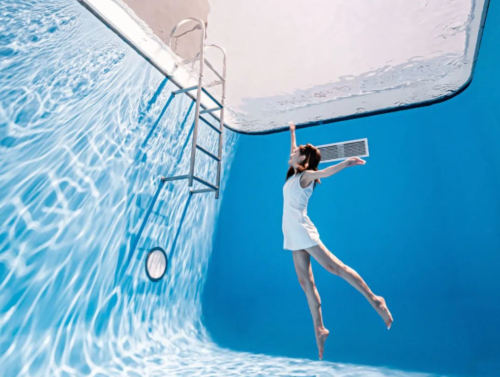 A woman in a white dress underwater in a swimming pool, near the pool ladder, with blue walls and reflections of water on the pool floor.