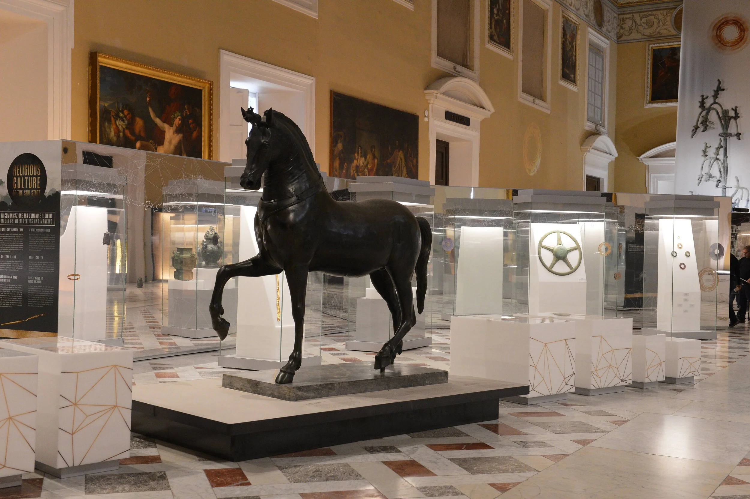 Indoor art exhibit featuring a large black horse sculpture at the center, surrounded by glass display cases with artifacts, ornate wall paintings, and decorative elements in a grand hall.