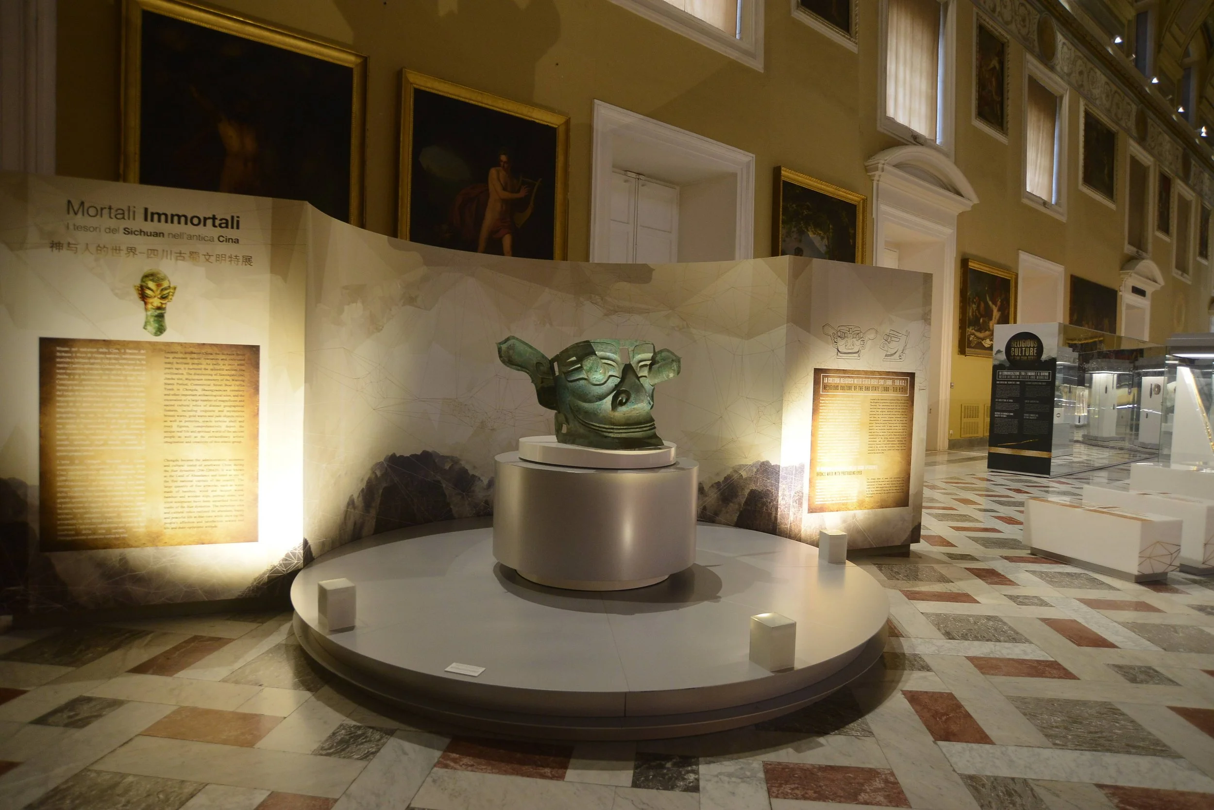 An ancient bronze mask displayed on a round pedestal in a museum exhibit. The background features informational panels and framed artwork on the wall.