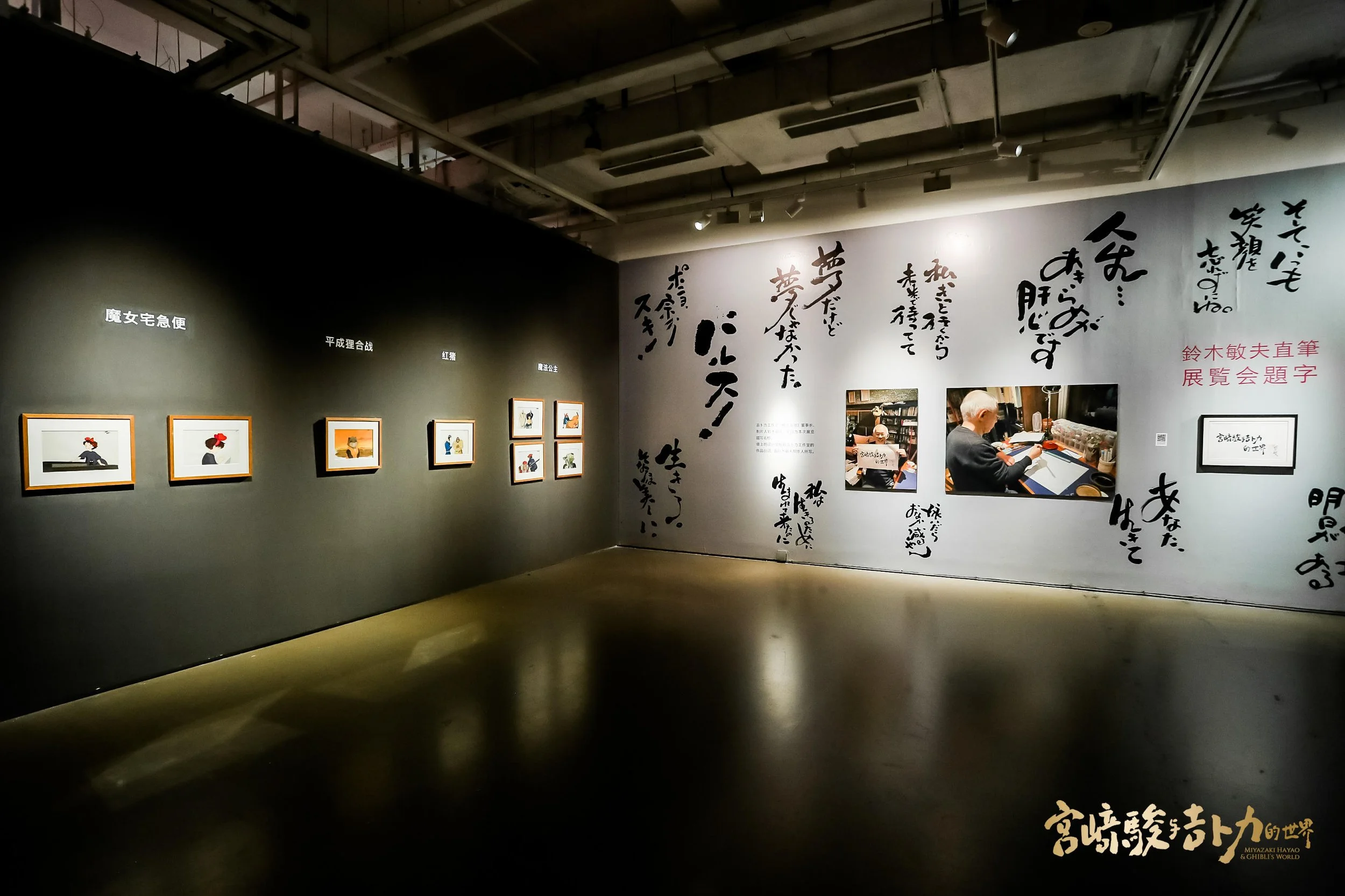 Museum exhibit of framed illustrations on a dark wall, large Japanese calligraphy on the adjacent wall, and photographs of an elderly man writing or drawing, in a dimly lit gallery space.