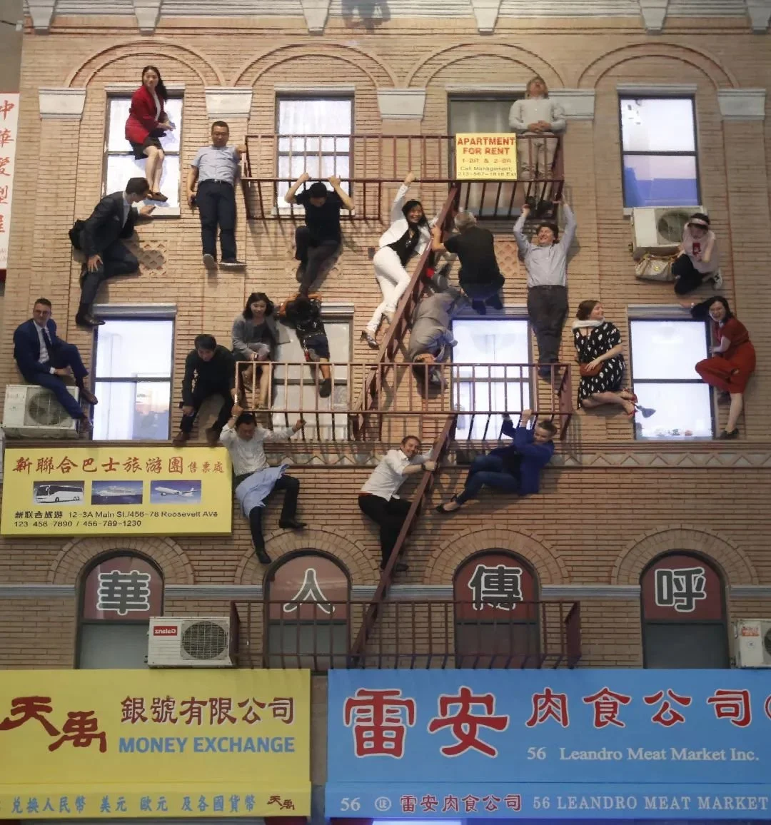 Employees are arranged on the exterior of a building to create an optical illusion of climbing, sitting, and hanging from the fire escape, with some posing like they are climbing or hanging.