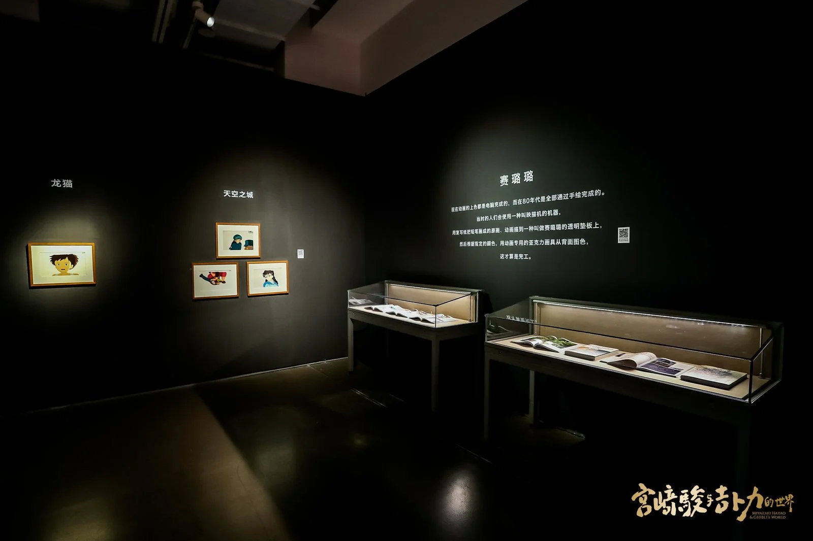 Dark exhibition room with four framed artwork on a black wall, three on the right and one on the left. Two glass display cases containing open books are positioned below a wall with text in Chinese. The room has focused lighting highlighting the artw