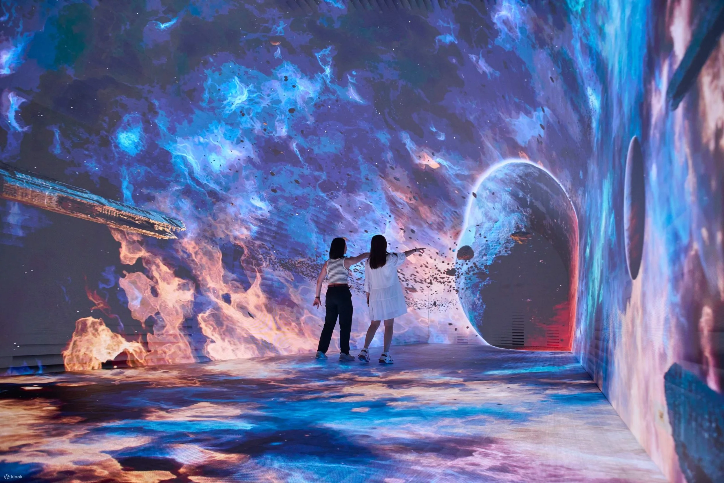 Two people standing inside an immersive digital art exhibit displaying a cosmic scene with planets, stars, and nebulae.