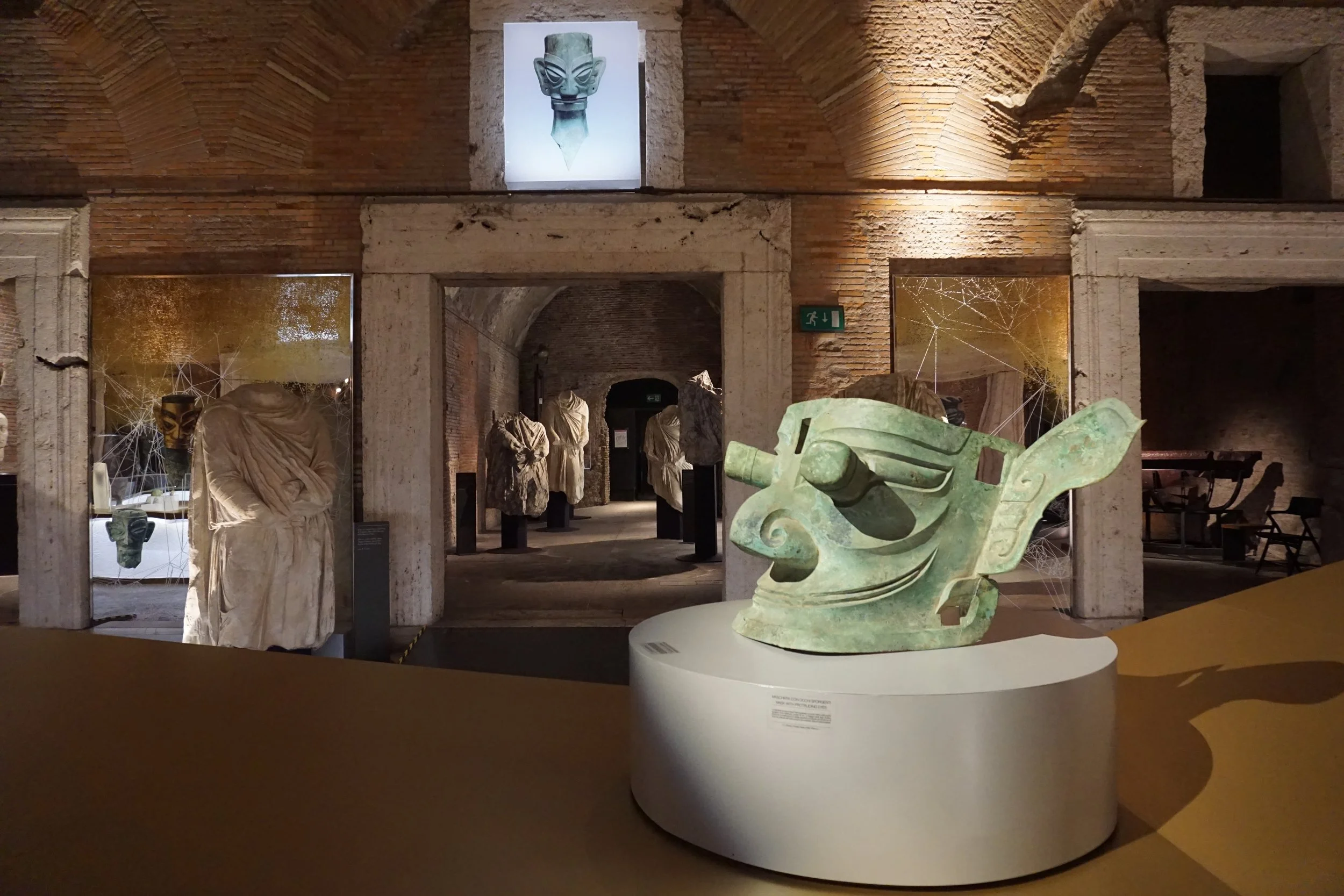 Ancient sculptures and artifacts displayed in a museum with brick walls, including a prominent green-colored sculpture of a stylized face or mask in the foreground, and various statues covered in cloth in the background.