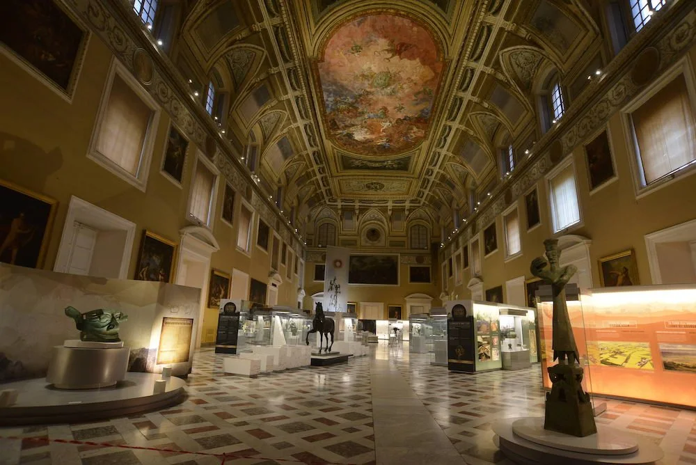 Interior of a museum with high ornate ceiling, paintings on the walls, statues, and display cases.