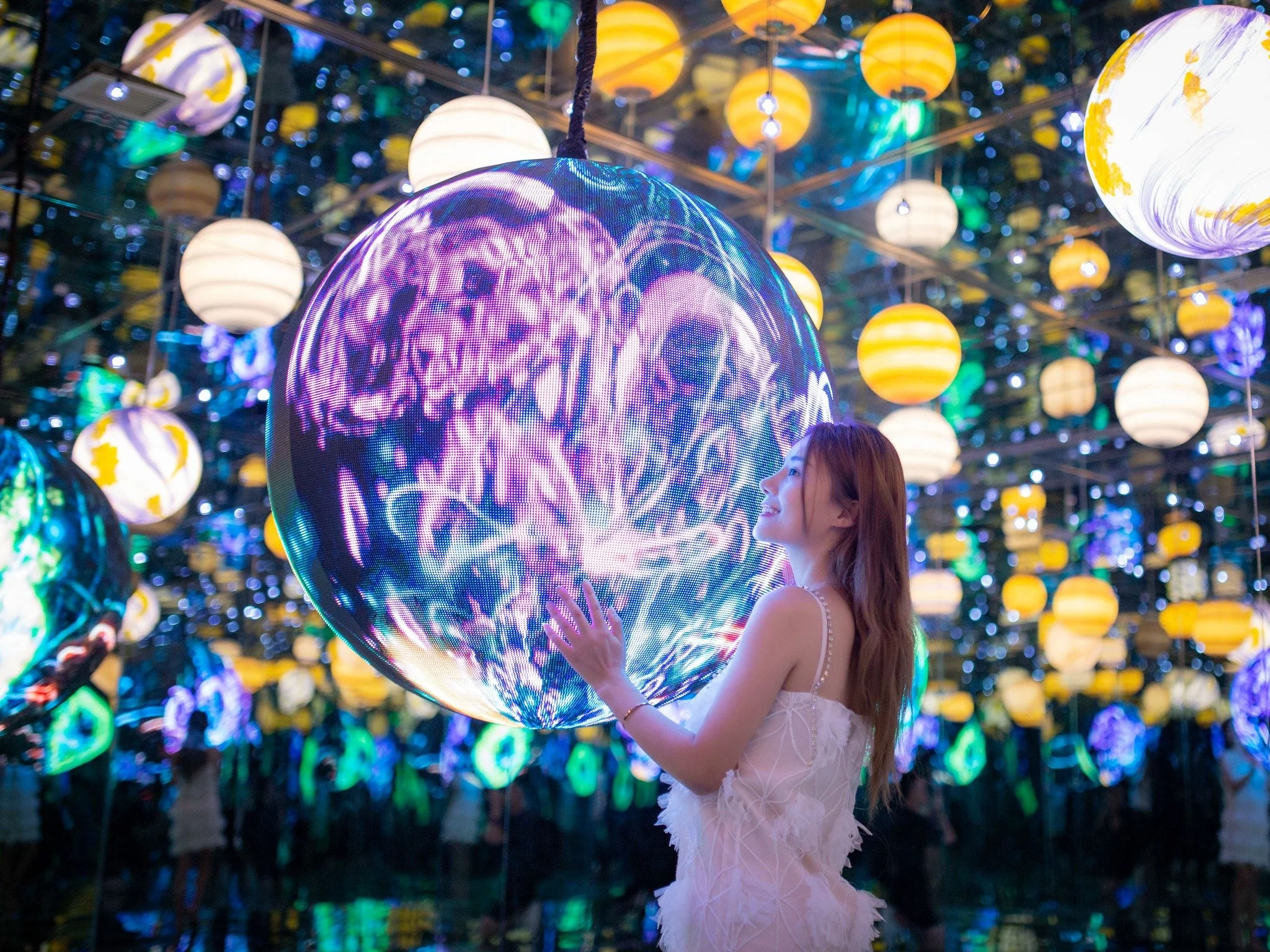 Young woman standing next to a large glowing, colorful LED sphere with intricate patterns, surrounded by hanging lanterns in a vibrant, illuminated art installation.