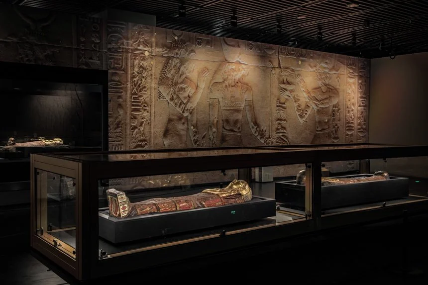 Ancient Egyptian artifacts displayed in glass cases with a large wall relief of Egyptian figures and hieroglyphics in the background.