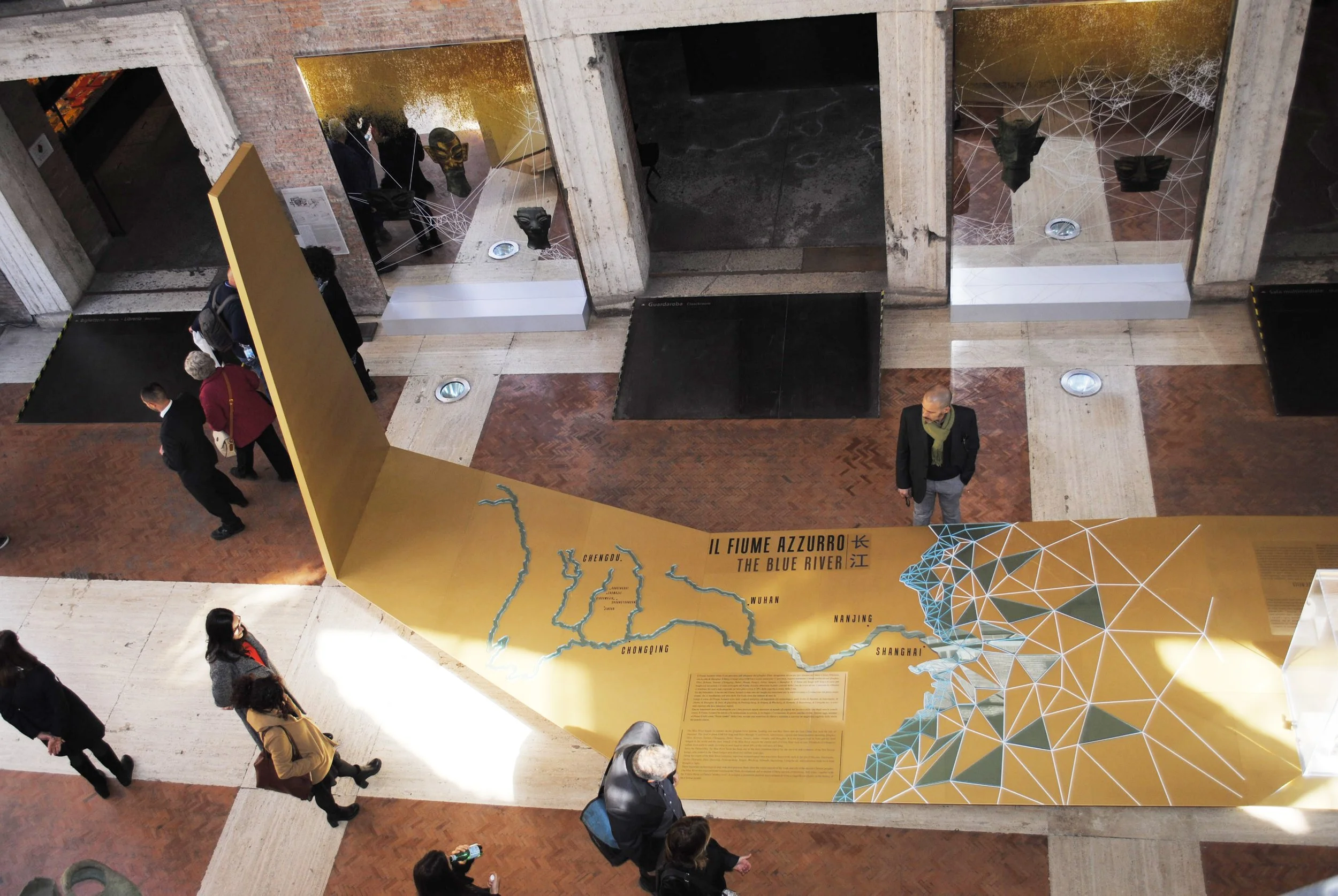An indoor exhibit showing a large, yellow, curved display board with a map of the Blue River in China. The display includes text in English and Chinese, and an illustrated geometric representation of the river's flow. Several visitors are walking aro